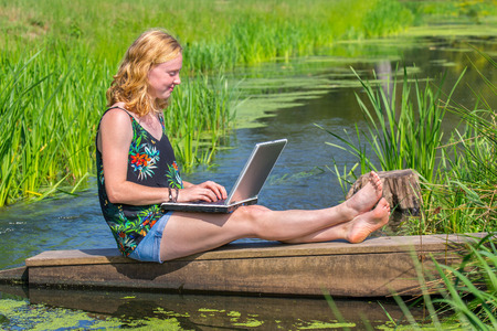 Young red-haired woman operating laptop near water in natureの写真素材