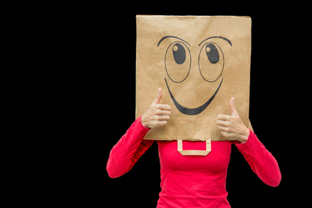 Woman wearing paper bag with happy face isolated on black. She is holding her thumbs upの写真素材
