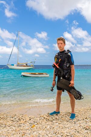 European male diver standing on beach of Bonaire with sea and boatsの写真素材