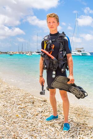 dutch male diver posing on beach of Bonaire with ocean and boatsの写真素材