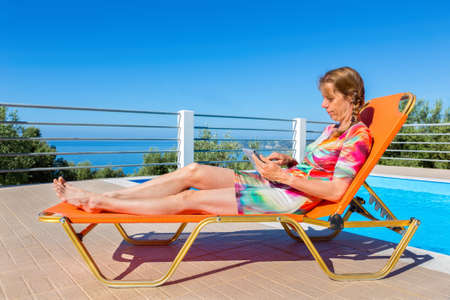 Middle aged dutch woman lying on orange sunlounger reading tablet near poolの写真素材