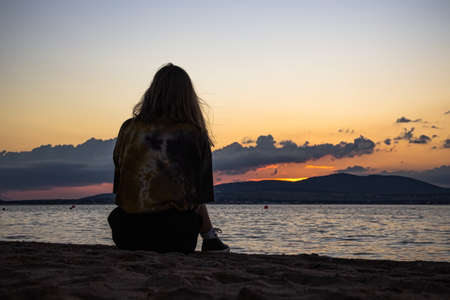 A young girl is sitting on a sandy beach, completely alone and watching the sunset. Dramatic summer...