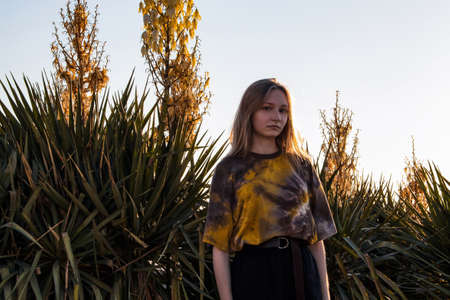 A young girl, blonde, in fashionable clothes, stands by a flowerbed next to a sandy beach, against...