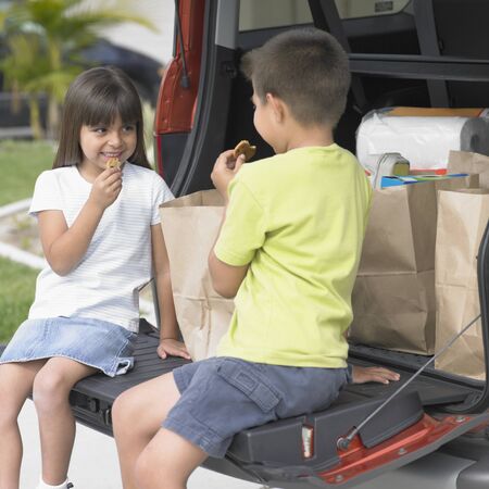 Brother and his sister sitting at the back of a car and eating biscuitsの写真素材