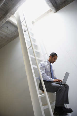 Businessman sitting on ladder with laptopの写真素材