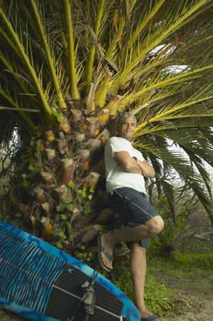 Senior man leaning against a palm tree next to a surfboard, Oakland, California, United Statesの写真素材