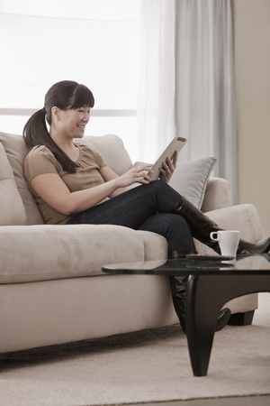 Asian woman sitting on sofa using digital tabletの写真素材
