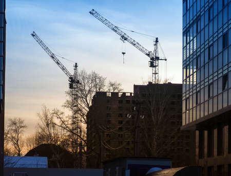 Two cranes raised their arrows over the unfinished multi-story building. Nearby is a residential building with a glass surface. Electric wires run along housesの写真素材