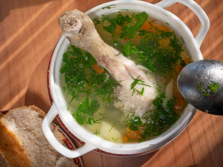 Appetizing soup with vegetables, herbs and chicken leg. Asterisks of fat float on the surface of the broth. A spoon lies on a bowl. On a plate is a piece of bread.の写真素材