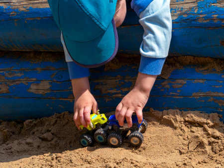 In the handles of a small child there are two children's toy cars of different colors, which are directed towards each other. The kid bent over the painted logs and rolls the cars on the sand.の写真素材