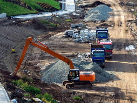 Landscaping, road asphalting. The excavator digs the ground. Dump trucks brought gravel. There are several piles of gravel and reinforced concrete blocks on the ground.の写真素材
