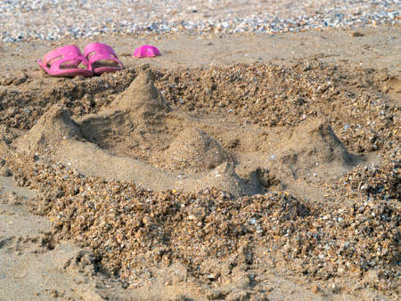 Children's sand castle on the seashore. There are flip flops next to it, and a sand mold.