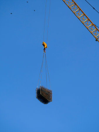 Tower crane lifts metal rebar. The load is suspended on a crane hook against a blue sky. Birds are flying in the sky.の写真素材