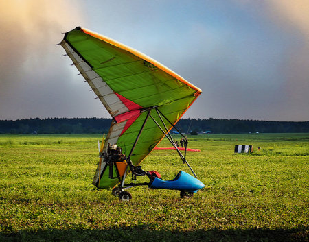 A hang glider with a motor stands on the grass in a field. There are people standing in the...