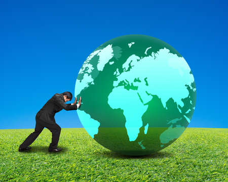 businessman rolling large green ball with global map on it with green meadow and clear blue sky backgroundの写真素材