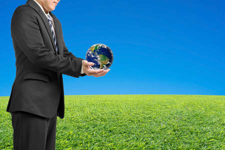 Businessman two hands hold ball with global map with fresh green meadow and clear blue skyの写真素材