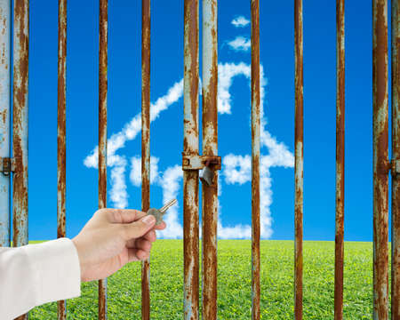 Hand hold key unlocking locked door with cloud house on green meadow in blue sky backgroundの写真素材
