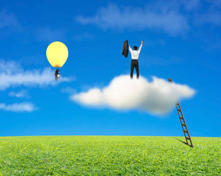 Businessman standing on cloud, another in lamp balloon with meadow and blue sky backgroundの写真素材