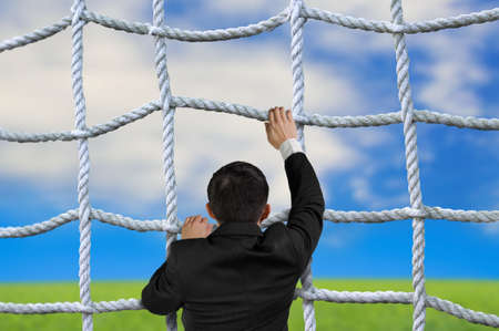 Businessman climbing the crisscross rope net on natural sky cloud grass backgroundの写真素材