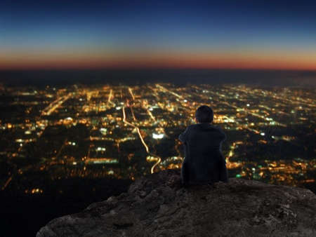Businessman sitting on rock mountain with city view at nightの写真素材