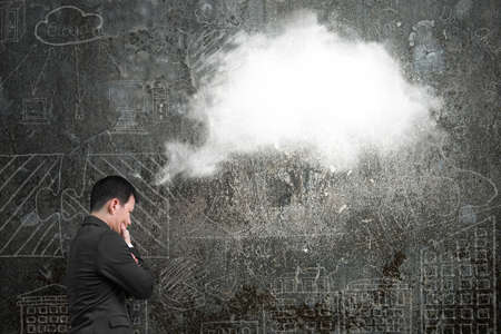Businessman thinking about white cloud thought bubble above his head with doodles wall backgroundの写真素材