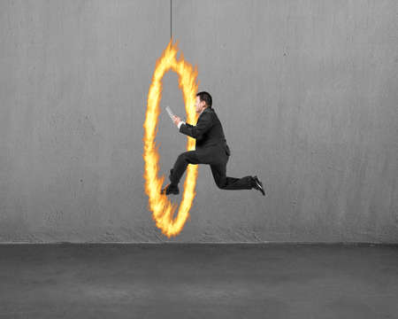 Businessman holding tablet jumping through fire hoop with concrete wall backgroundの写真素材