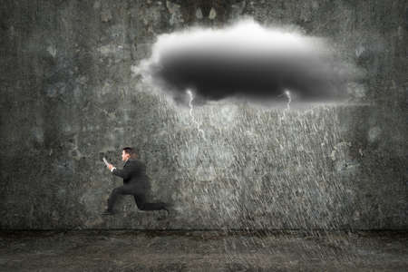 Running businessman holding tablet with dark clouds raining and lightning on concrete wall backgroundの写真素材