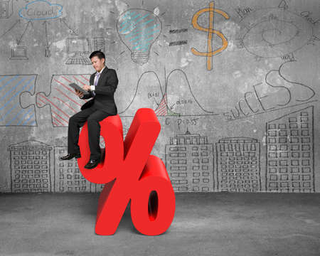 Using tablet businessman sitting on red percentage sign with business concept doodles wall backgroundの写真素材