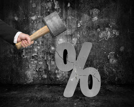 Hand holding sledgehammer hitting cracked percentage sign with dark concrete room backgroundの写真素材