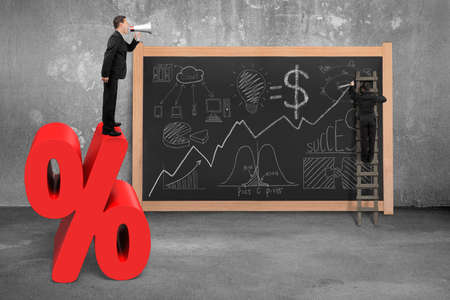 Businessman using megaphone standing on red percentage sign with business concept doodles and concrete room blackboardの写真素材