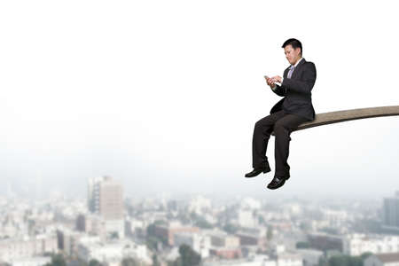 Using mobile phone businessman sitting on springboard with cityscape backgroundの写真素材