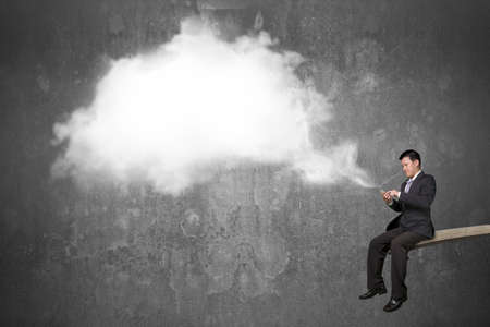 Using mobile phone businessman sitting on springboard with white cloud thought bubble and concrete...