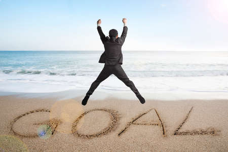 Jumping businessman cheering for goal word written on sand beach background