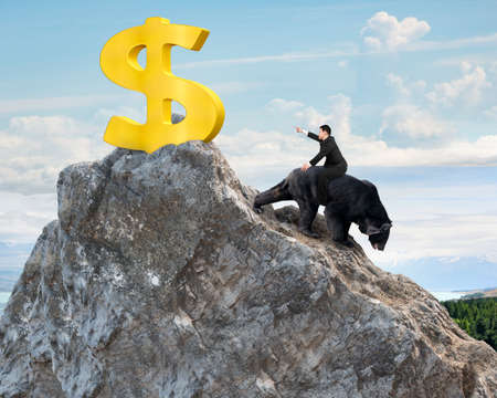 Businessman riding black bear pursuing gold dollar sign on mountain peak with sky clouds background. Fight back bearish market concept.の写真素材