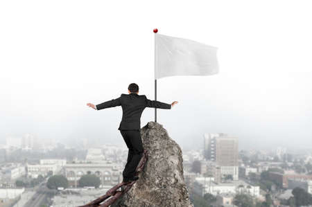 Businessman walking and balancing on old iron chain toward white flag of mountain peak with cityscape backgroundの写真素材