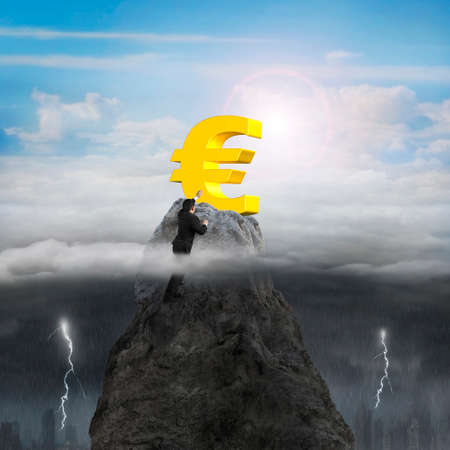 Businessman hand wanting for golden euro symbol on mountain peak high in the sky with opposite weather conditions background, sunny sky cloudscape, dark overcast lightning raining cityscapeの写真素材