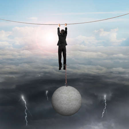 Businessman shackled by heavy concrete ball hanging on the rope with opposite weather conditions sky background, bright sun cloudscape and dark cloudy lightning.の写真素材