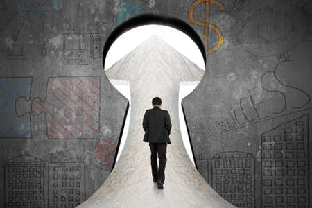 Businessman walking on marble road toward keyhole door with business concept doodles concrete wall backgroundの写真素材