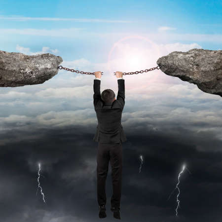 Businessman hand hanging on the cracking rusty iron chains connect two cliffs with opposite weather conditions sky background, bright sun cloudscape and dark cloudy lightning.の写真素材