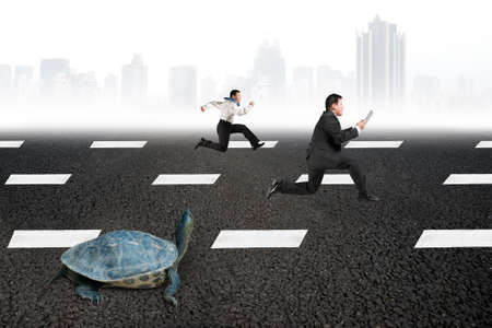 Two businessmen running with turtle on asphalt road with gray urban scene skyline background. Tortoise race competing metaphor concept.の写真素材