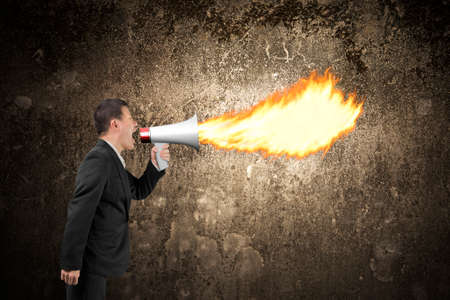 Angry businessman screaming into megaphone spitting fire flame illuminated the dark mottled concrete wall backgroundの写真素材