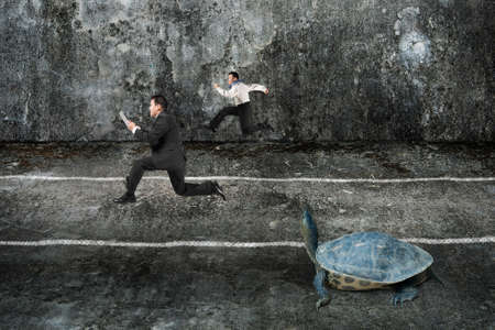 Two businessmen running with turtle in the dark mottled concrete room. Tortoise race competing metaphor concept.の写真素材