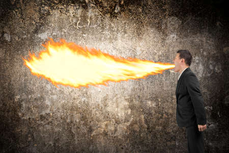 Angry businessman spitting fire flame from mouth illuminated the dark mottled concrete wall backgroundの写真素材