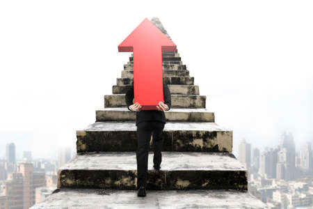 Businessman carrying big 3D red arrow sign and climbing on old concrete stairs with urban scene backgroundの写真素材