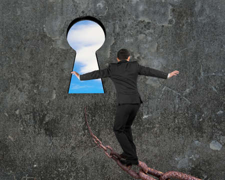 Man balancing on old iron chain toward keyhole, with sky clouds view and gray concrete wall...