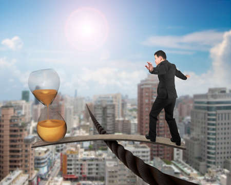 Businessman and golden hourglass on wood board, balancing on wire, with sky sun cityscape background.の写真素材