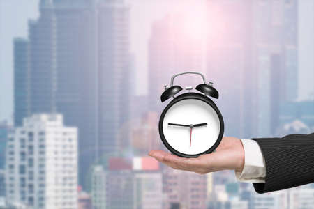 Hand holding alarm clock with sunlight city background.