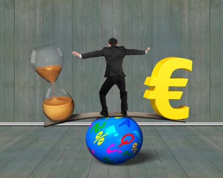 Man standing between hourglass and golden euro sign, balancing on seesaw of wood board and colorful ball, with wooden wall and floor indoors background. Time is money concept.の写真素材