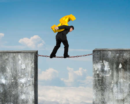 Businessman carrying golden dollar sign balancing on rusty chain connected two high dirty concrete walls, with blue sky clouds background.の写真素材