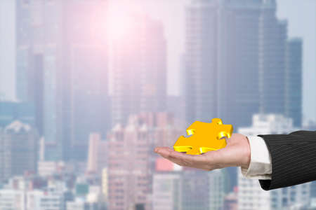 Male hand holding one 3D gold jigsaw puzzle piece, with sunlight city background.の写真素材